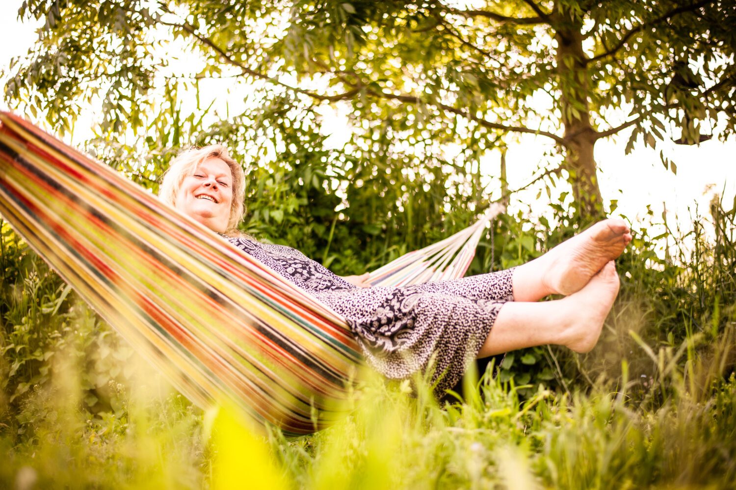 Een vrouw zit speels in een hangmat. ze draagt een kleedje met bloemen en heeft blote voeten.