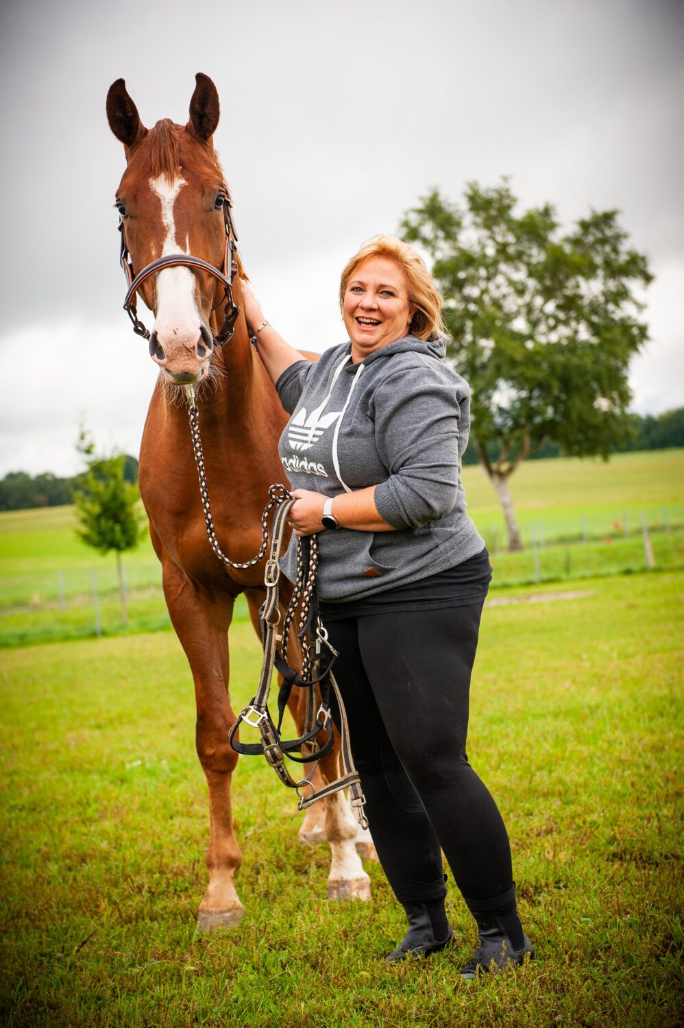 Een vrouw staat lachend naast een paard in de weide. Ze heeft een hand op de hals van het paard, en de andere hand houdt een touw vast.