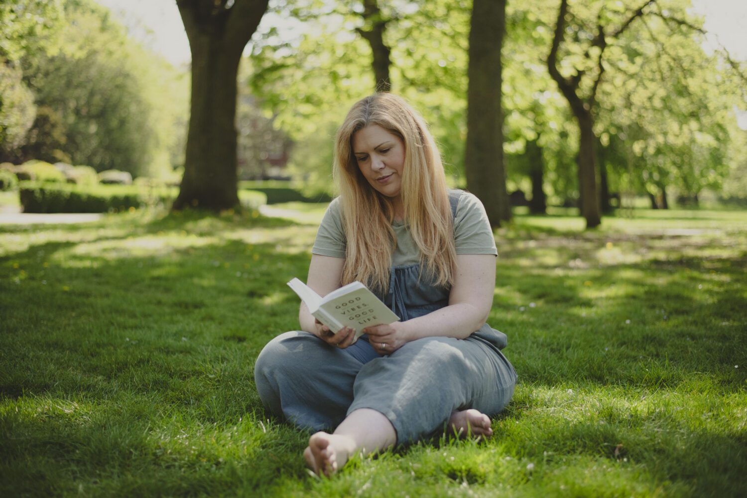 Een jonge vrouw zit in kleermakerszit een boek te lezen in het gras in een park.